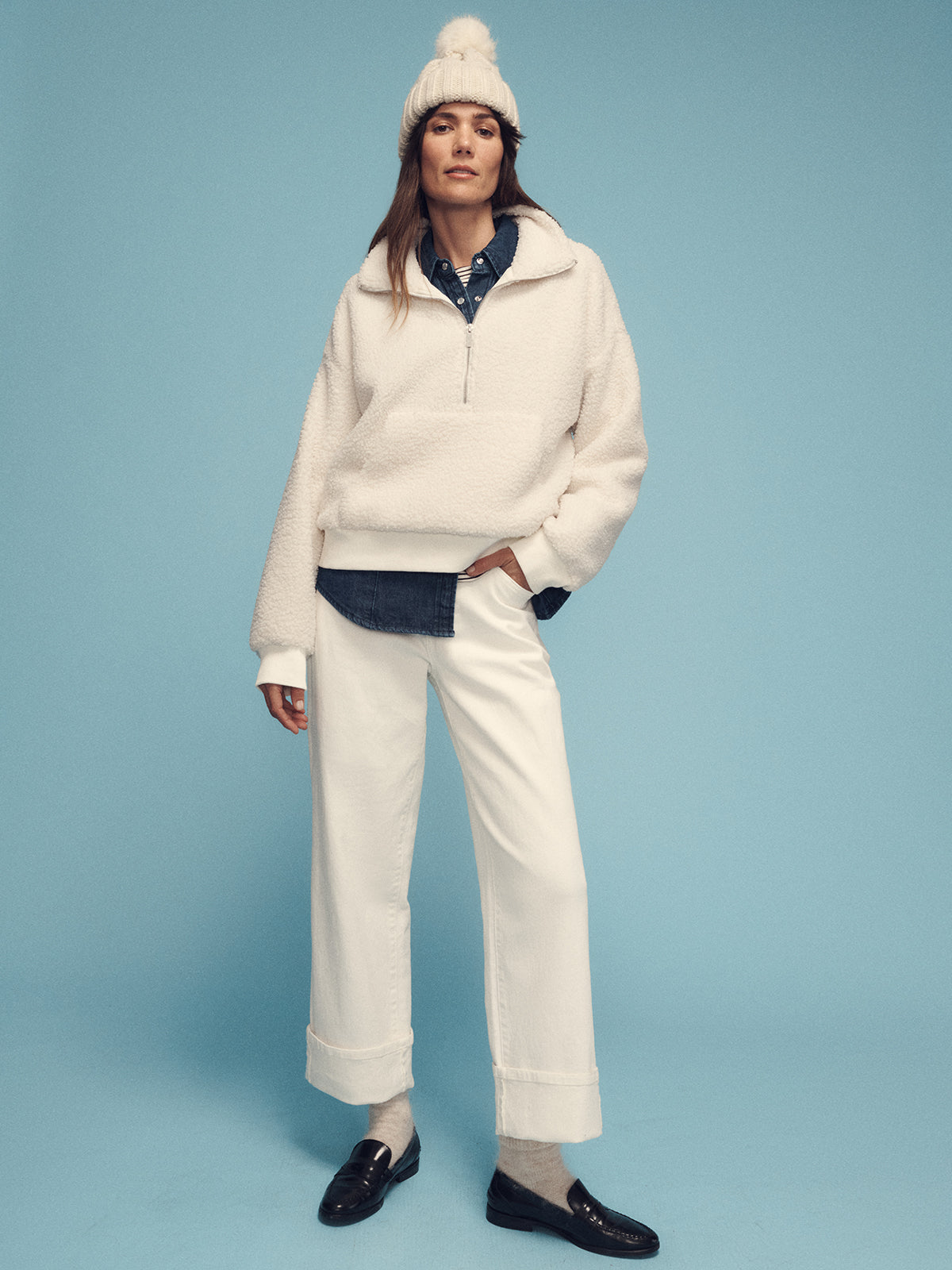 A person stands against a blue background wearing Sanctuary Clothing's alpine sherpa half zip popover in chalk, paired with white pants and a denim shirt underneath. They're accessorized with a knitted hat featuring a pom-pom and black loafers, their hand casually tucked in their pocket to exude a warm, relaxed style.