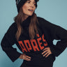 A person wearing the Sanctuary Clothing après intarsia logo sweater in black and mars red leans slightly forward against a blue background. They are also wearing black ski goggles with reflective lenses on a black knit hat with a pom-pom.