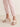 A person is standing on a light wooden floor, wearing light pink cuffed pants and the Brisk Sandal in Nude Bronze Sand Dollar by Sanctuary Clothing. The open-toe heels, featuring wide straps and a chunky, textured design, draw attention to the lower legs and feet.