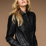 A woman with long, wavy blonde hair is wearing the Davidson Trucker Jacket Black by Sanctuary Clothing. She has her hands in her pockets and is standing against a brown background, looking slightly away from the camera with a neutral expression.