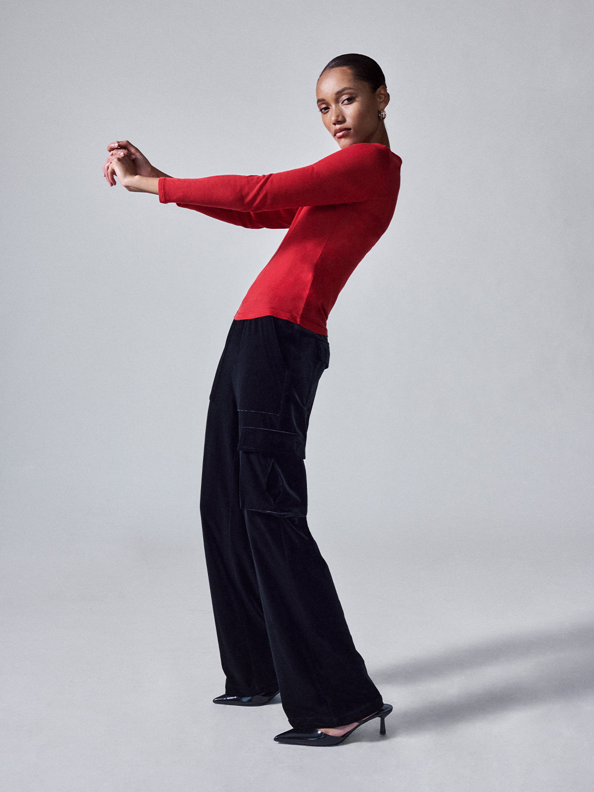 A woman in a Sanctuary Clothing Relaxed Velvet Reissue Black top and black wide-leg pants strikes a dynamic pose against a plain grey background. She leans back with her arms bent and extends her legs, wearing pointed black heels.