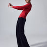 A woman in a Sanctuary Clothing Relaxed Velvet Reissue Black top and black wide-leg pants strikes a dynamic pose against a plain grey background. She leans back with her arms bent and extends her legs, wearing pointed black heels.