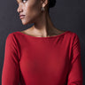 A person stands against a dark background, wearing a Sanctuary Clothing Semi Sheer Tee in Cherry Red and silver hoop earrings. They are looking to the left with an expression of calm contemplation, their hair neatly pulled back in a bun.