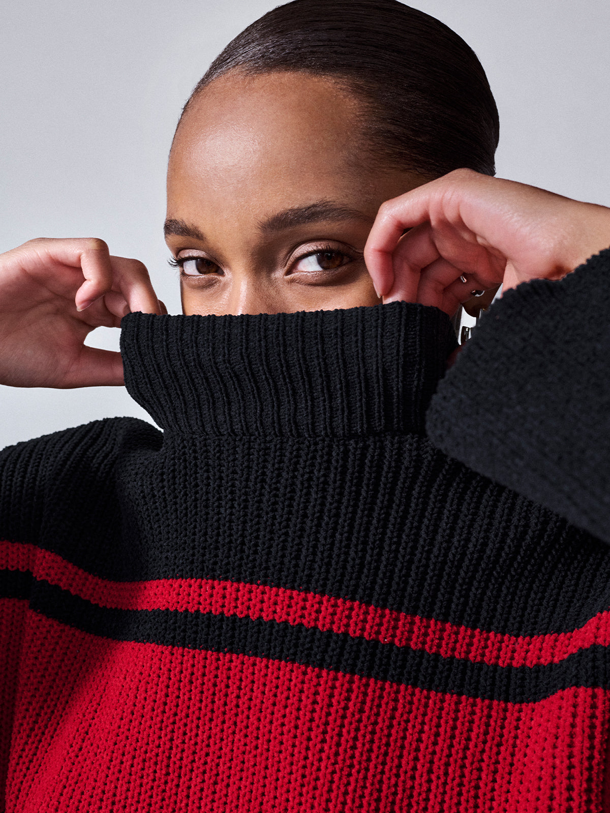 A person with dark, pulled-back hair is partially covering their face with the high collar of the Stay Cozy Sweater Black Red Stripe by Sanctuary Clothing. They are looking directly at the camera, with hands raised to adjust the collar. The background is plain and light-colored.