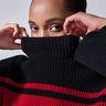 A person with dark, pulled-back hair is partially covering their face with the high collar of the Stay Cozy Sweater Black Red Stripe by Sanctuary Clothing. They are looking directly at the camera, with hands raised to adjust the collar. The background is plain and light-colored.