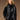 A woman with long blonde hair, wearing the Ryder Moto Jacket Black by Sanctuary Clothing over a black turtleneck, stands against a brown background. She is looking to the side with her hands in her pockets.