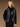 A woman with long blonde hair, wearing the Ryder Moto Jacket Black by Sanctuary Clothing over a black turtleneck, stands against a brown background. She is looking to the side with her hands in her pockets.