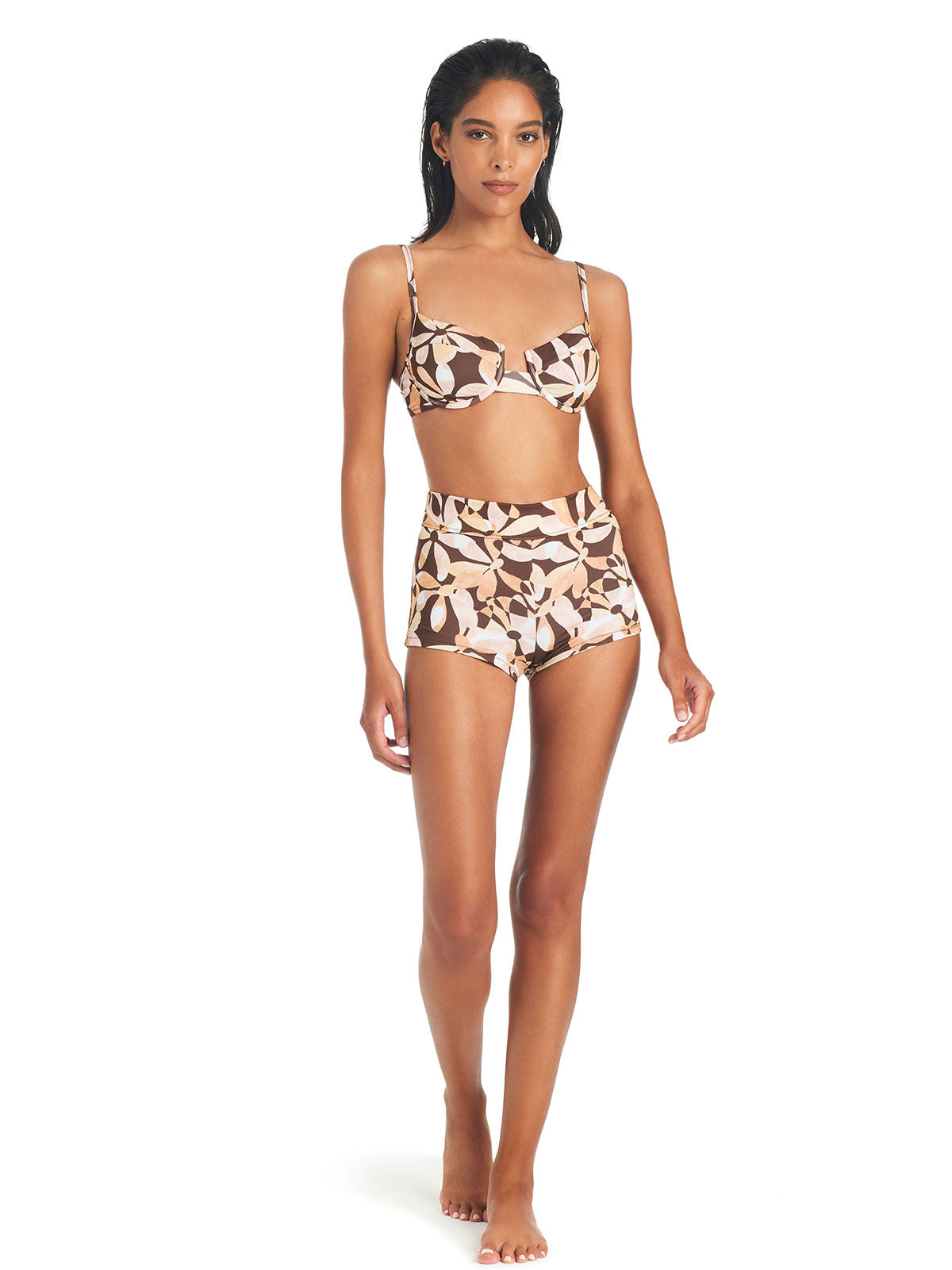 A woman with long dark hair poses in the Jigsaw Petals Bikini Top Chocolate by Sanctuary Clothing. The bikini features large floral designs in light and dark earth tones. She stands on a white background, showcasing both the front of the bikini top and bottoms.