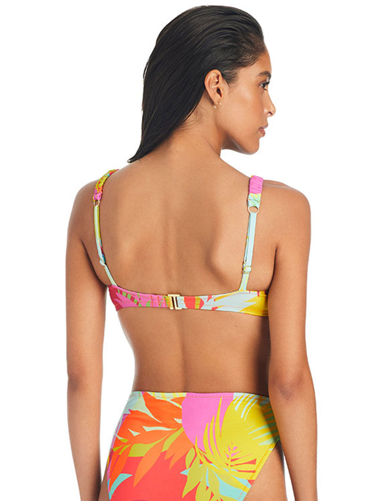 A woman is seen from the back wearing Sanctuary Clothing’s Palm Hottie Bikini Top Multi Palm Hottie, featuring a vibrant tropical leaf pattern in shades of pink, yellow, orange, and turquoise. She has dark, smooth hair and stands against a plain white background.