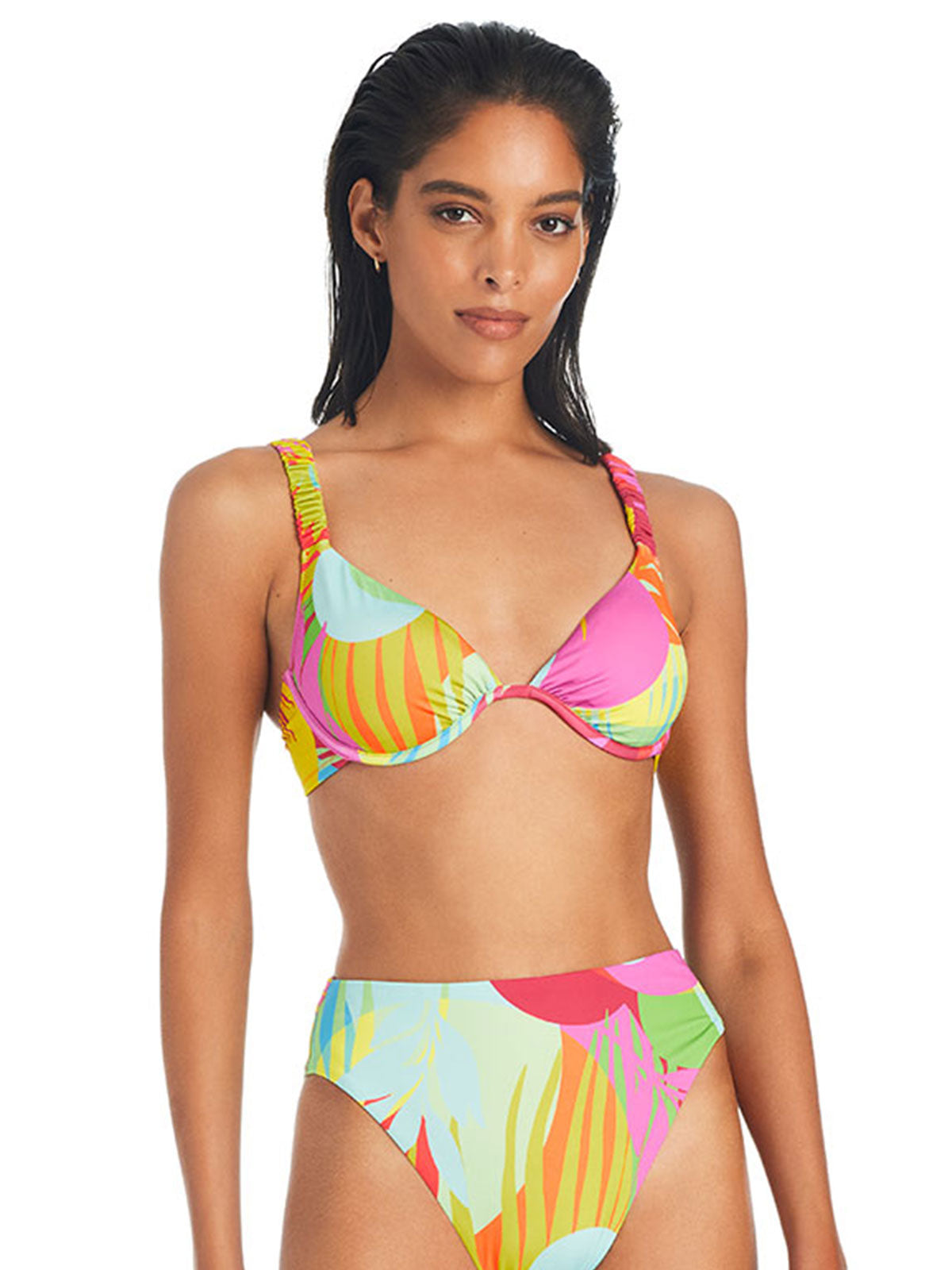 A woman is standing and modeling the Palm Hottie Bikini Top Multi Palm Hottie by Sanctuary Clothing. The bikini top boasts supportive straps, while the matching high-waisted bottoms share the same vibrant tropical design. She has wet, swept-back hair and a neutral, confident expression.