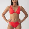 A person with long hair is wearing the Refresh Rib Double Layer Underwire Bikini Top in Mars Red from Sanctuary Clothing, standing against a plain, light-colored background. They carry a confident expression with their hands on their hips.