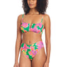 A woman with curly hair is wearing the vibrant Super Bloom V Wire Bandeau Bikini Top Multi by Sanctuary Clothing. The bikini top features a green base adorned with vibrant pink and yellow floral patterns. She stands with a relaxed posture, looking slightly to the left.