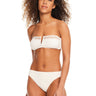 A woman with curly hair is wearing the Sea Crochet Hipster Bikini Bottom in White Sand from Sanctuary Clothing, paired with a matching strapless top featuring a small V-shaped cut-out in the center. She is standing against a plain white background.