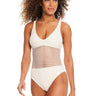 A woman with curly hair smiles while wearing the Sanctuary Clothing Sea Crochet Scoop Neck One Piece Swimsuit in White Sand, featuring a net-like, see-through midsection. She is standing against a plain white background.