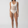 Dressed in the Sanctuary Clothing's sea crochet high leg scoop neck one piece in white sand, a person stands self-assuredly. They pair the cream-hued swimsuit, featuring a mesh panel midsection, with black and white sandals against a plain, light-colored backdrop that highlights the swimwear.