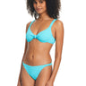 A person with curly hair is smiling while modeling the Sandbar Solids Hipster Bikini Bottom in Curacao from Sanctuary Clothing. The light blue swimsuit features a knotted detail on the top and matching bikini bottoms. The individual stands in a relaxed pose against a plain white background.