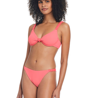 A woman with curly hair is wearing a watermelon sugar bikini from Sanctuary Clothing's Sandbar Solids collection. She is standing and posing with one hand resting by her side. The bikini includes a top with a knotted center and simple bottoms. The background is white.