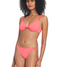 A woman with curly hair is wearing a watermelon sugar bikini from Sanctuary Clothing's Sandbar Solids collection. She is standing and posing with one hand resting by her side. The bikini includes a top with a knotted center and simple bottoms. The background is white.