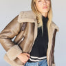 A person with long blonde hair wears the Zarina Shearling Coat in Mink Beige from Sanctuary Clothing over a black sweater and blue jeans. They pose confidently against a neutral background.