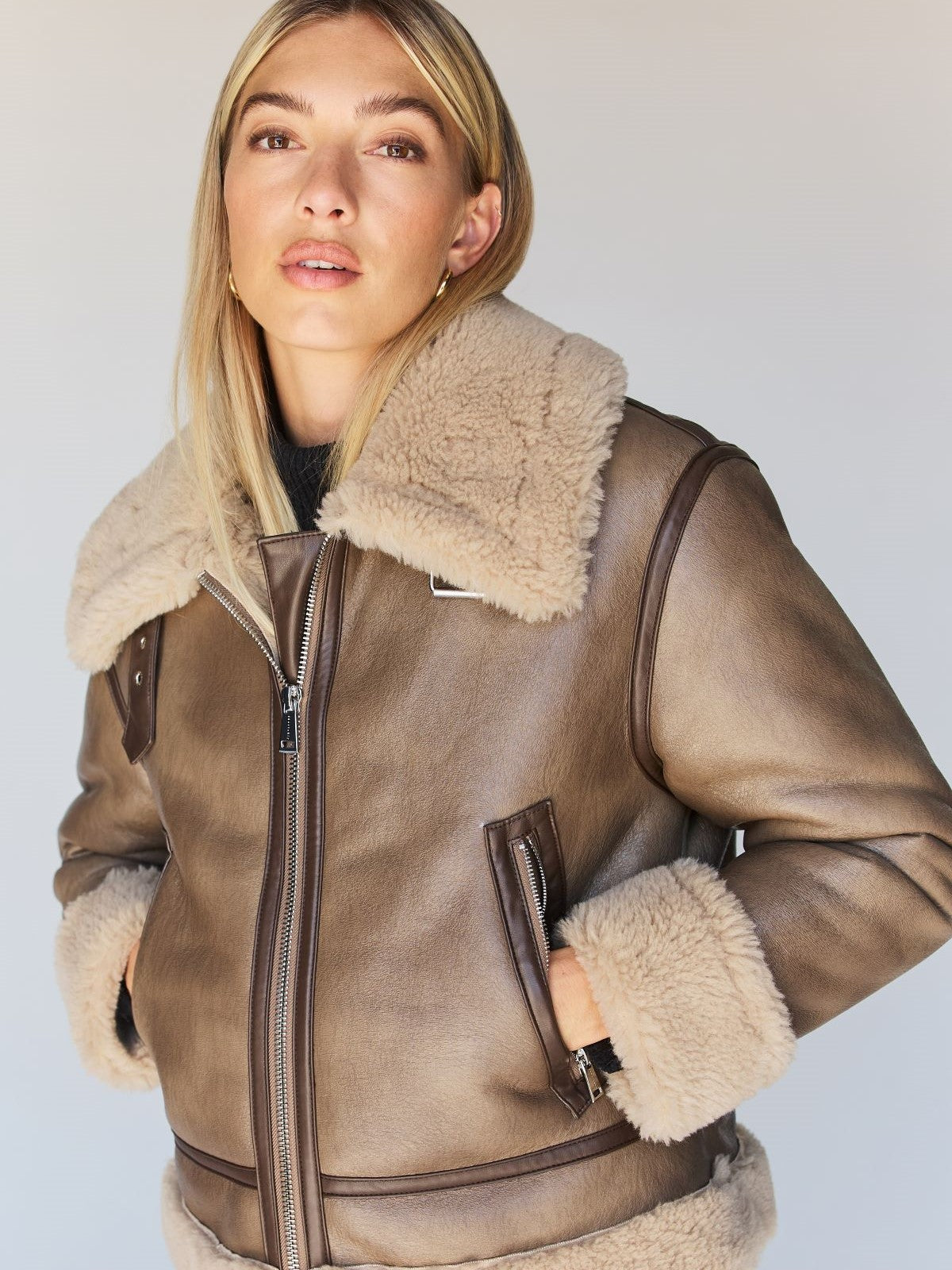 A person with long blonde hair wearing the Zarina Shearling Coat Mink Beige from Sanctuary Clothing, looking at the camera. The coat features a large, fluffy collar and cuffs, with a zipped front. The background is plain and light-colored.