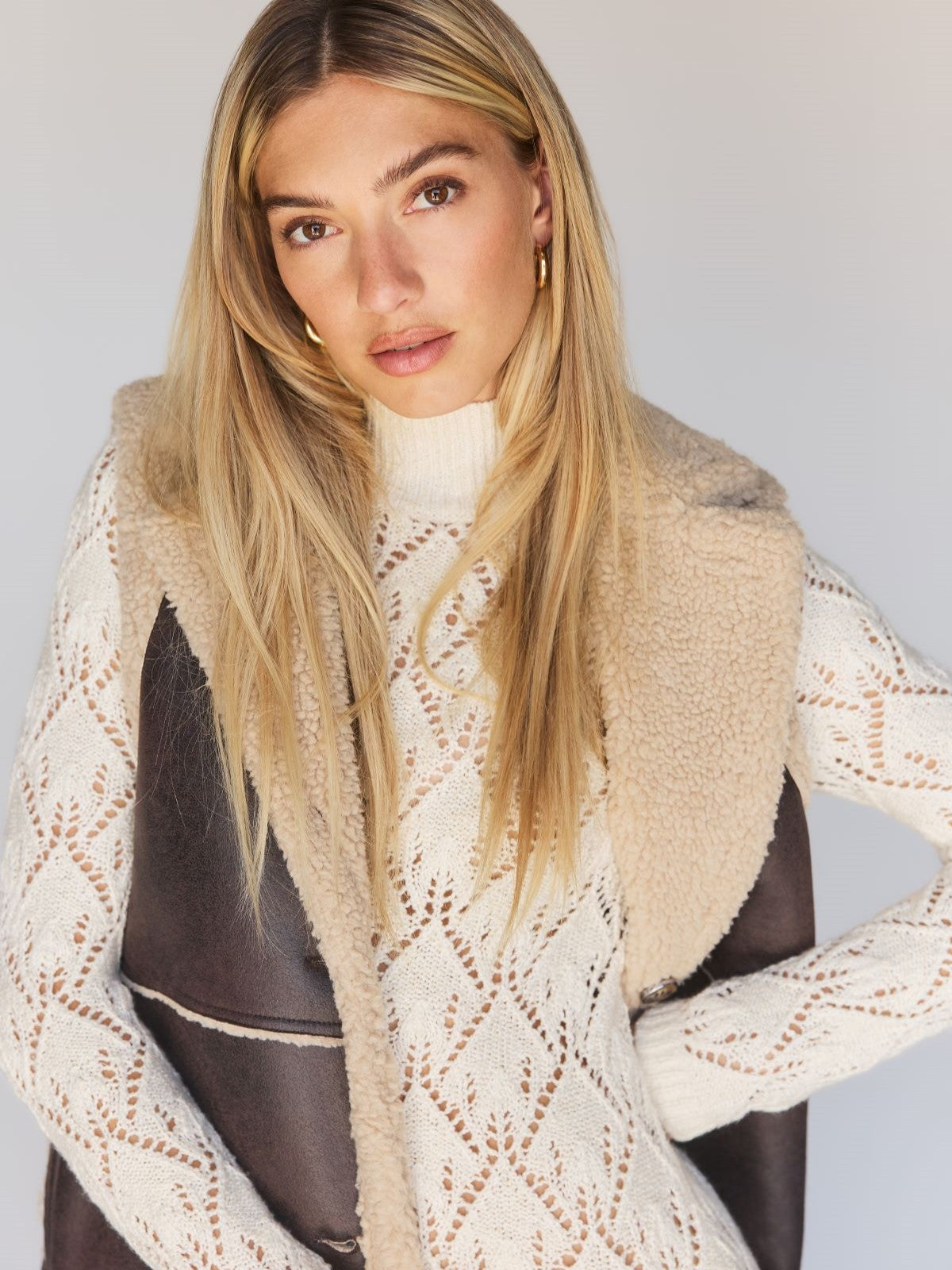 A woman with long blonde hair poses confidently, wearing a cream-colored knit sweater with a patterned design and the Paisley Shearling Vest in Espresso from Sanctuary Clothing. She accessorizes with gold hoop earrings against a neutral backdrop.