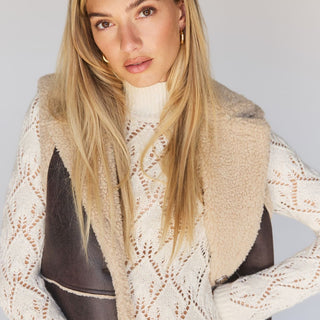 A woman with long blonde hair poses confidently, wearing a cream-colored knit sweater with a patterned design and the Paisley Shearling Vest in Espresso from Sanctuary Clothing. She accessorizes with gold hoop earrings against a neutral backdrop.