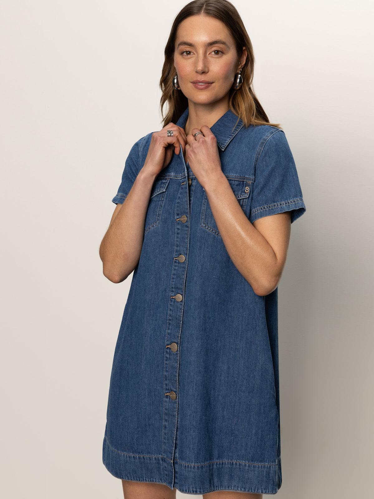 A woman poses against a plain backdrop wearing the Sanctuary Clothing denim shiftdress in medium wash, featuring short sleeves, a button front, collar, and chest pocket. She touches her collar and smiles softly at the camera.