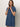 A woman poses against a plain backdrop wearing the Sanctuary Clothing denim shiftdress in medium wash, featuring short sleeves, a button front, collar, and chest pocket. She touches her collar and smiles softly at the camera.