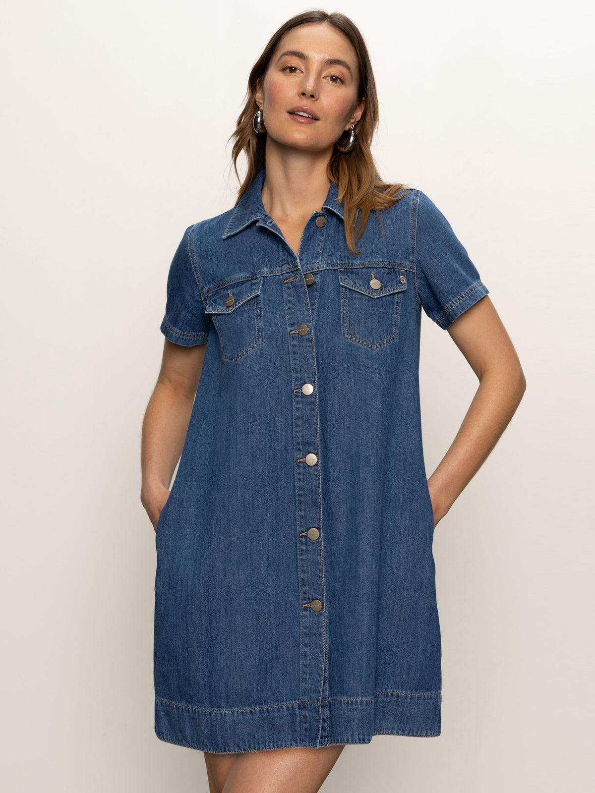 A woman stands against a light background wearing the Sanctuary Clothing denim shiftdress in medium wash, featuring short sleeves, a button front, chest pockets, and her hands in the side pockets.