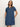 A woman stands against a light background wearing the Sanctuary Clothing denim shiftdress in medium wash, featuring short sleeves, a button front, chest pockets, and her hands in the side pockets.