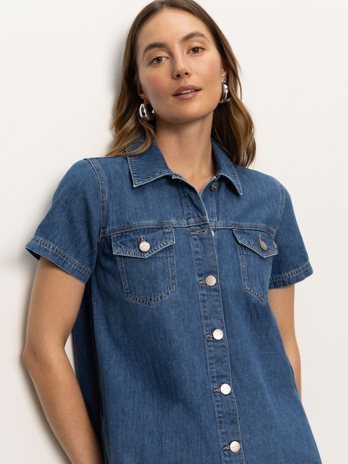 A woman with long brown hair, silver hoop earrings, and a neutral expression wears the Sanctuary Clothing denim shiftdress in medium wash, standing against a light background and looking at the camera.