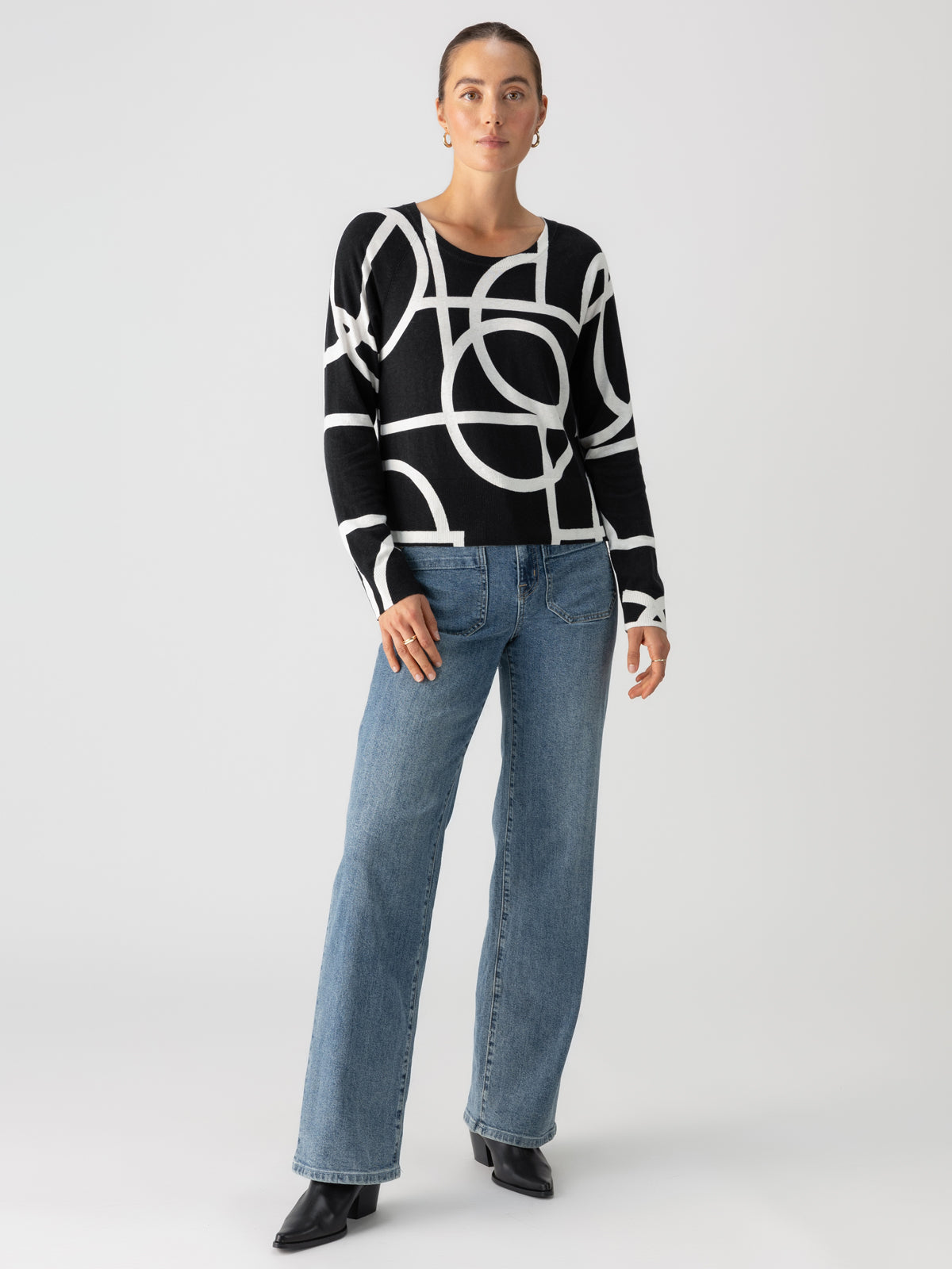 A person with hair pulled back stands in front of a white background. They are wearing the "All Day Long Sweater Graphic Lines Black" by Sanctuary Clothing, paired with blue jeans and black shoes. One hand is resting on their hip, and the other hangs by their side.