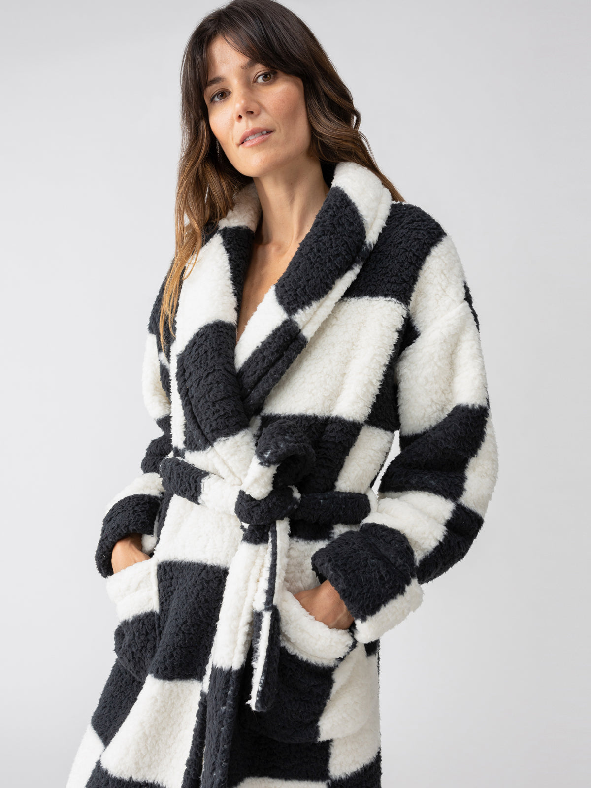 A woman wearing the Shawl Collar Teddy Robe in Black Check by Sanctuary Clothing, featuring a black and white pattern with pockets, stands against a neutral background.