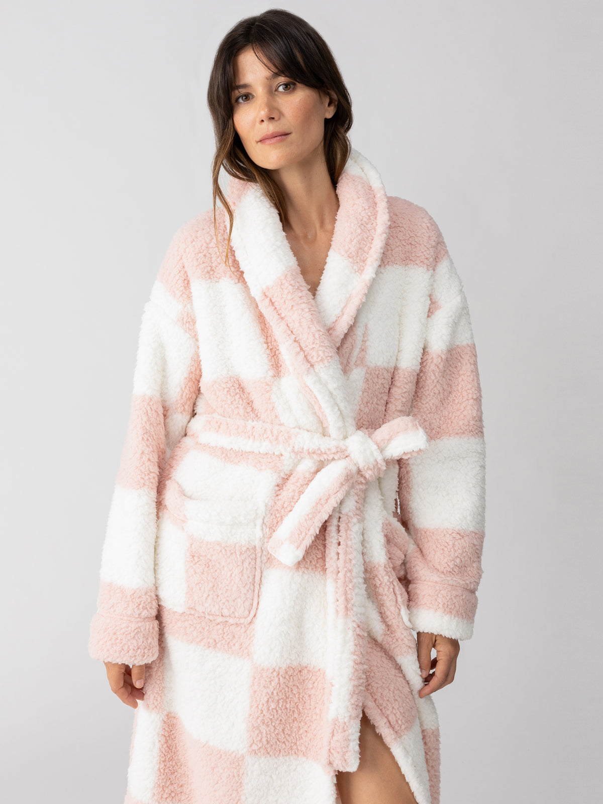 A person is wearing the Sanctuary Clothing shawl collar teddy robe in a rose check pattern, featuring pink and white squares. The robe includes a tied belt and pockets, and the individual has long, dark hair against a plain light-colored background.