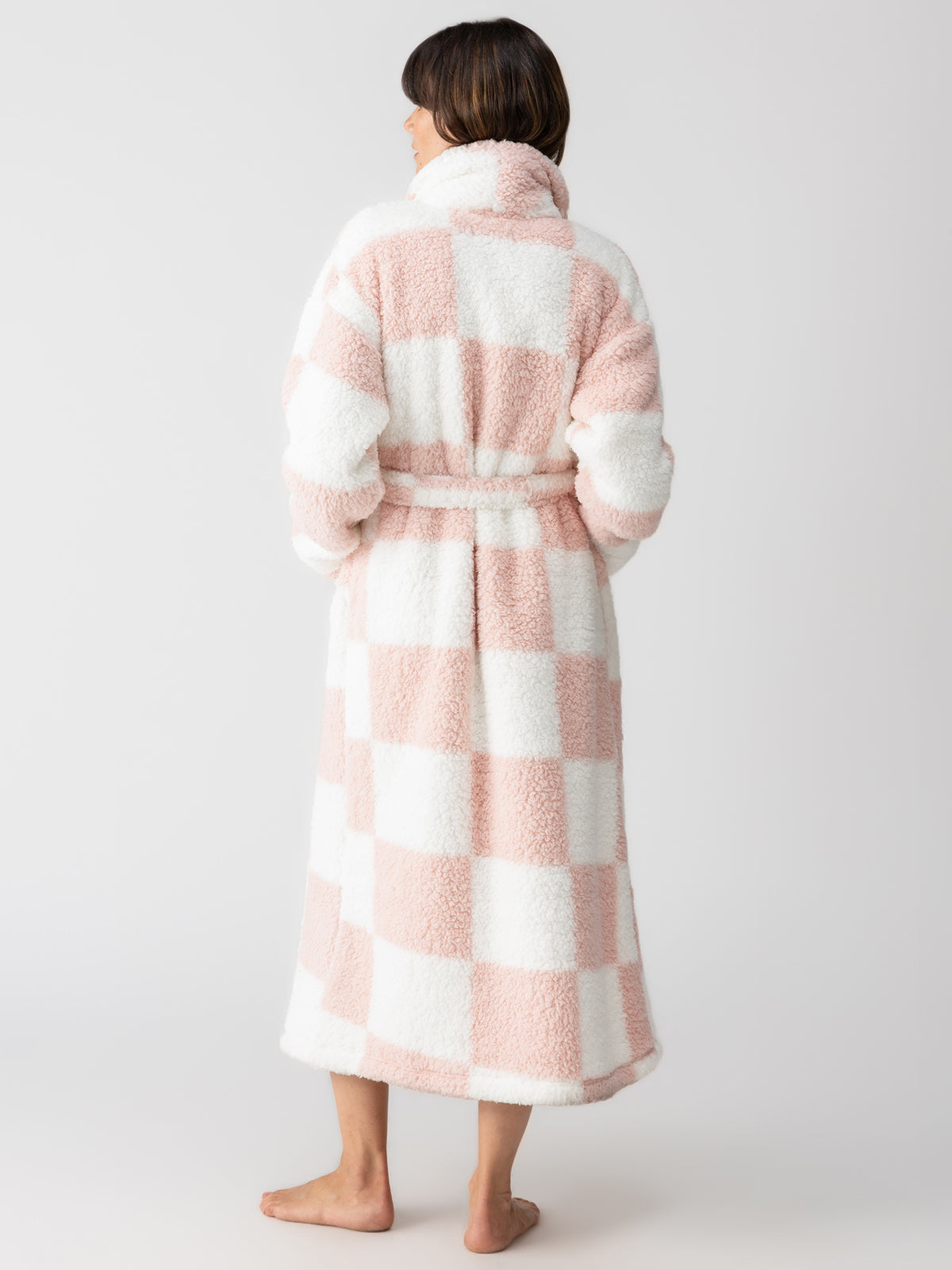 A person stands barefoot, facing away, wearing the Sanctuary Clothing shawl collar teddy robe in rose check. The fluffy robe is belted at the waist against a plain and light-colored background.