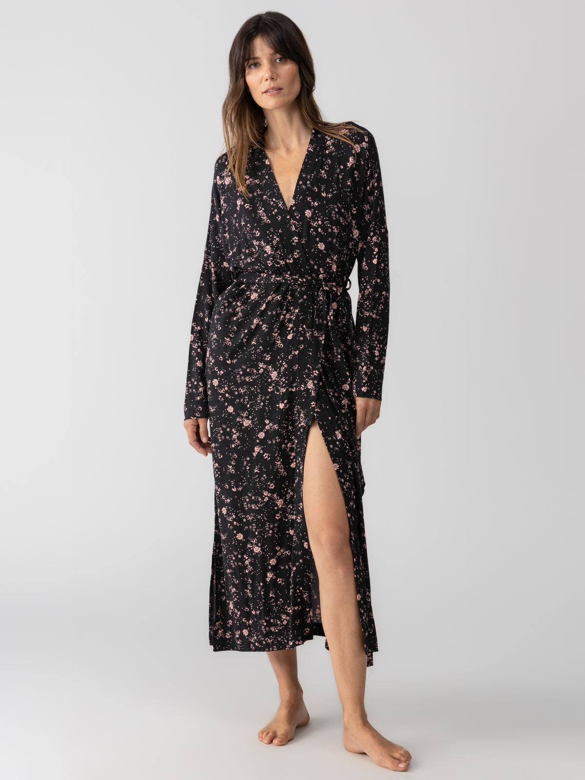 A barefoot woman wearing Sanctuary Clothing's Maxi Cardi Falling Flowers, a long black floral wrap dress with a high slit, stands against a plain white background. She has long, wavy hair and is posing with her left hand on her hip and her right arm relaxed by her side.