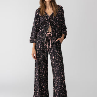 A woman stands barefoot against a plain background, wearing the Lounge Set Falling Flowers from Sanctuary Clothing, a matching black pajama set adorned with a floral pattern. The top features long sleeves and is partially unbuttoned, while the pants include an adjustable drawstring waistband. She has a relaxed expression.