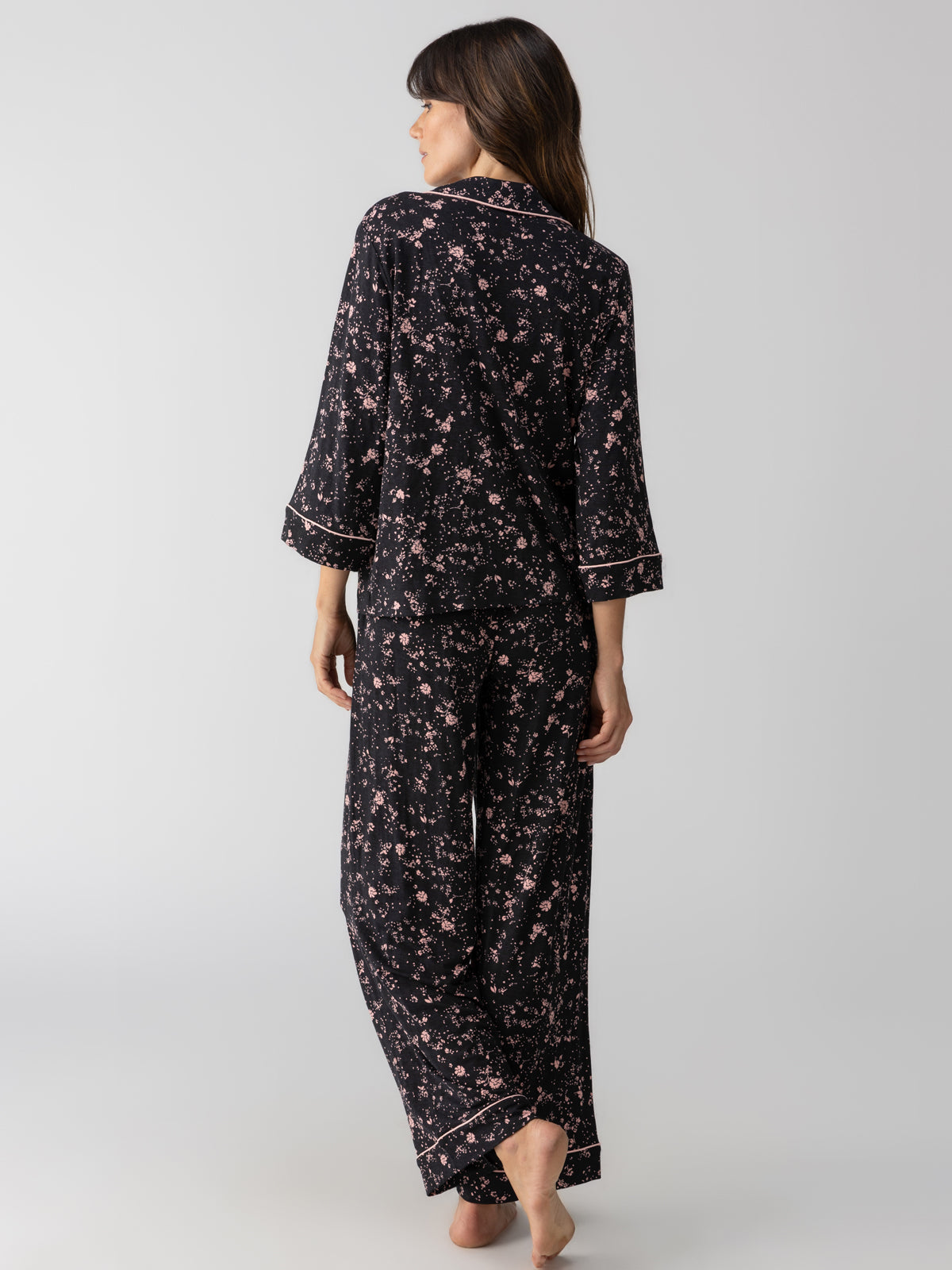 A person with long dark hair is standing barefoot, facing away from the camera. They are wearing the Lounge Set Falling Flowers by Sanctuary Clothing, featuring long sleeves and wide-leg pants adorned with a dark floral pattern. The background is plain and light-colored.