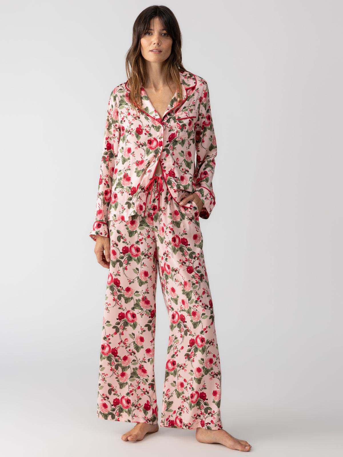 A person stands barefoot wearing the Sanctuary Clothing's "notch and pant set winter rose," featuring a pink floral design with red and green flowers. The outfit consists of a long-sleeved button-up top paired with matching wide-legged pants, all set against a plain white background.