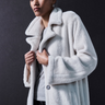 In a dark setting, someone confidently poses for the camera, wearing Sanctuary Clothing's "Live In Fur Coat Frosted White" over a gray top, with the coat buttoned and their hands grasping the lapels.