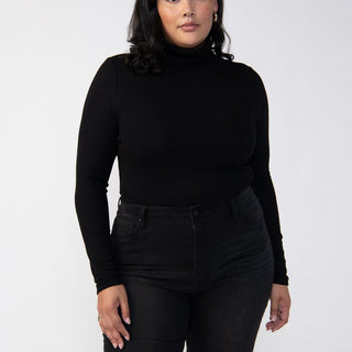 A person with medium-length dark hair is wearing the Essential Turtleneck Black from the Sanctuary Clothing Inclusive Collection and black jeans. They stand against a plain white background, looking directly at the camera with a confident and composed expression.