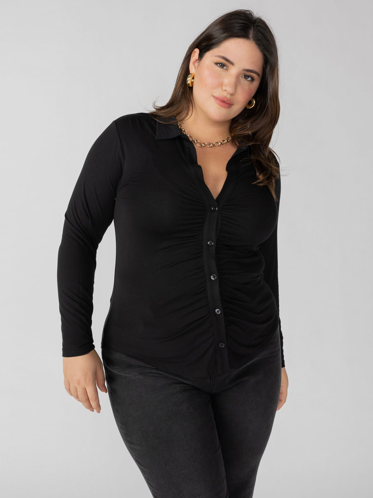 A woman with long dark hair wears the Dreamgirl Button Up Black Inclusive Collection blouse with long sleeves and matching black pants from Sanctuary Clothing. She is accessorized with gold jewelry, including a necklace and earrings, and stands against a plain, light grey background.