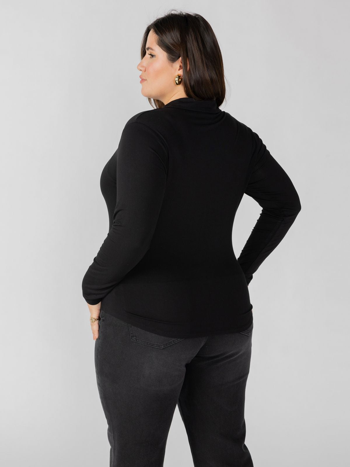 A woman with short dark hair is standing and facing away from the camera, wearing the Dreamgirl Button Up Black Inclusive Collection top by Sanctuary Clothing paired with dark jeans. The plain white background highlights her chic ensemble.