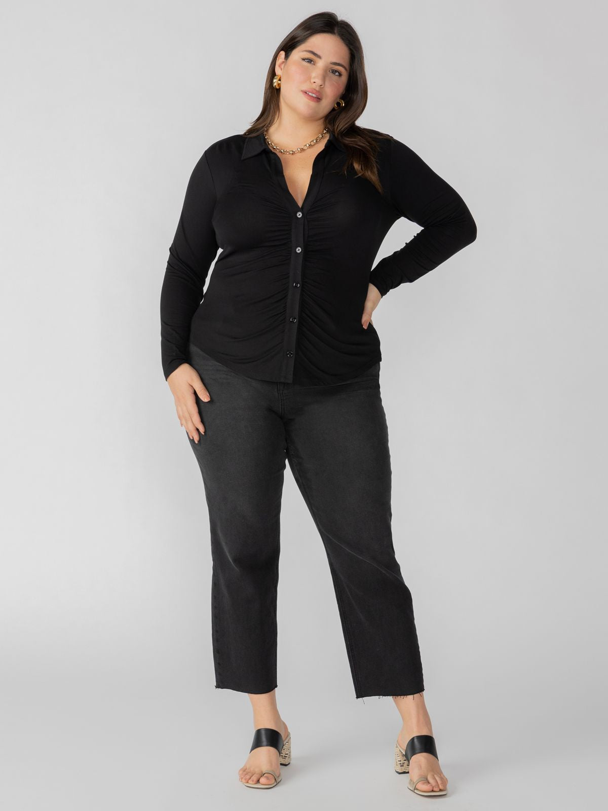 A woman stands confidently wearing the Dreamgirl Button Up Black from Sanctuary Clothing's Inclusive Collection, paired with black jeans. She has long, dark hair and is accessorized with gold earrings and a matching necklace. She is standing against a simple, light grey background and is wearing black, open-toed sandals.