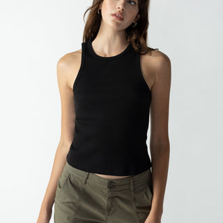 A person with long hair is wearing a black Ribbed Tank Top from Sanctuary Clothing and olive green cargo pants. They are standing against a plain, light gray background. The individual has a relaxed expression, with their head slightly tilted and their arms at their sides.