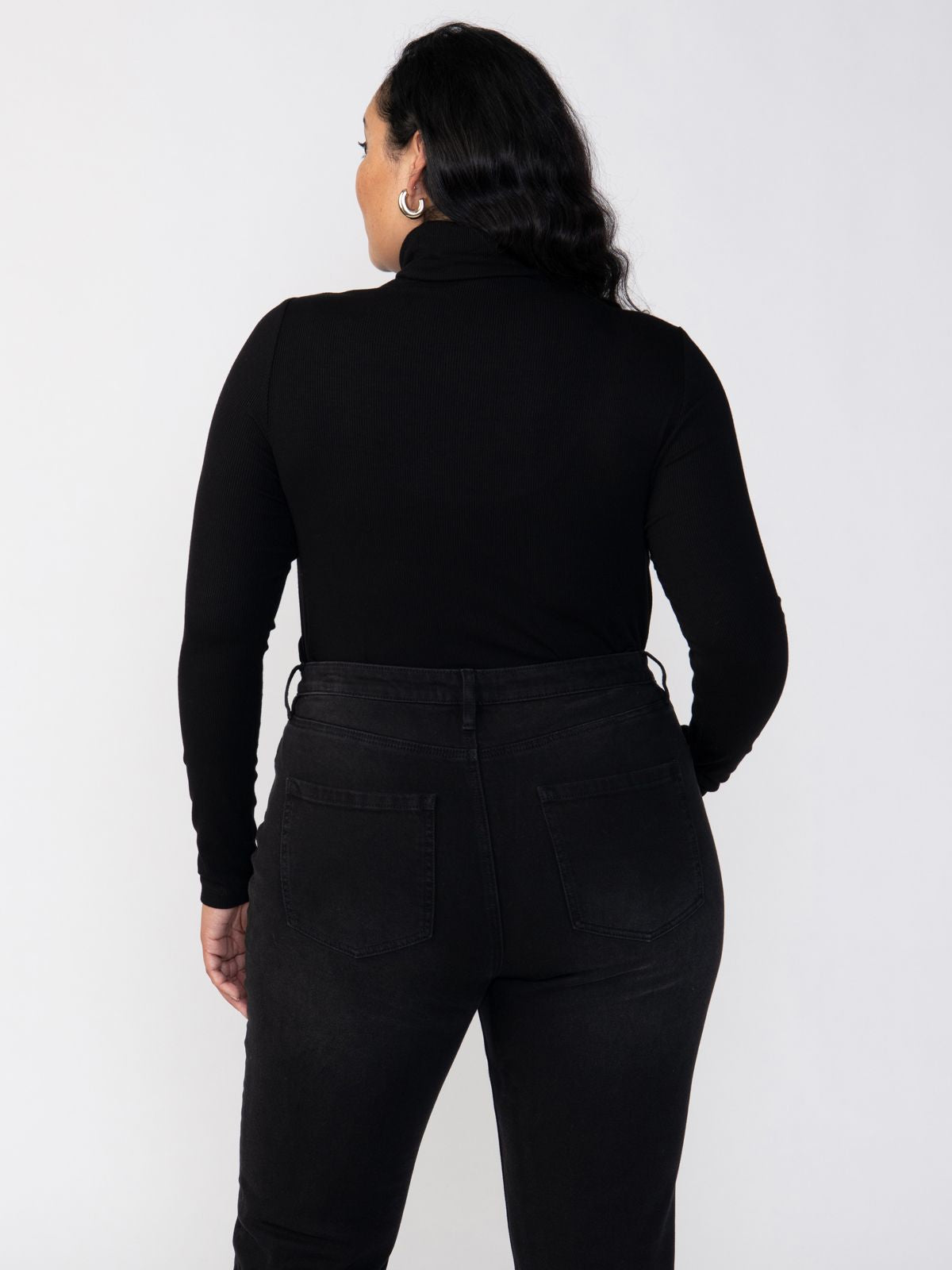 A person with long, wavy hair is standing with their back to the camera. They are wearing a Sanctuary Clothing Essential Turtleneck Black Inclusive Collection long-sleeve shirt and high-waisted black jeans. The background is a simple, light gray color.