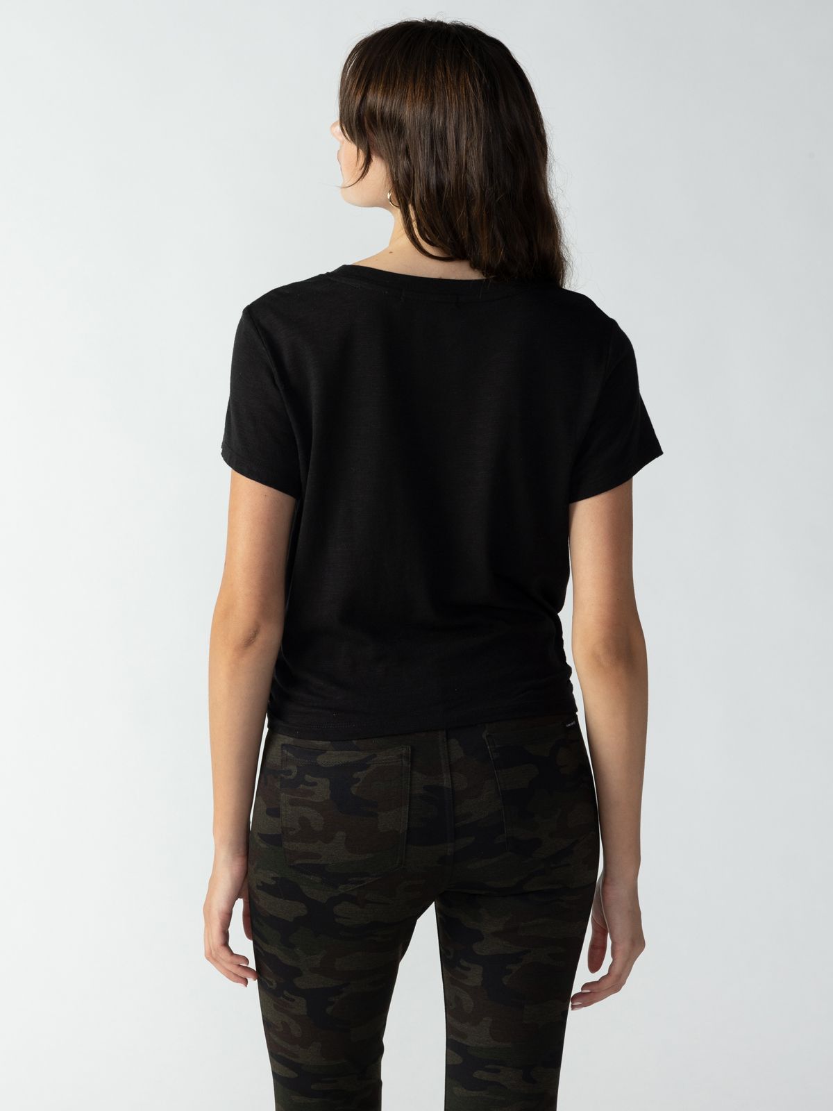 A person with long dark hair is standing facing away from the camera, wearing a Sanctuary Clothing Highstreet Tee in black and dark green camo-patterned pants. The background is plain and light-colored.